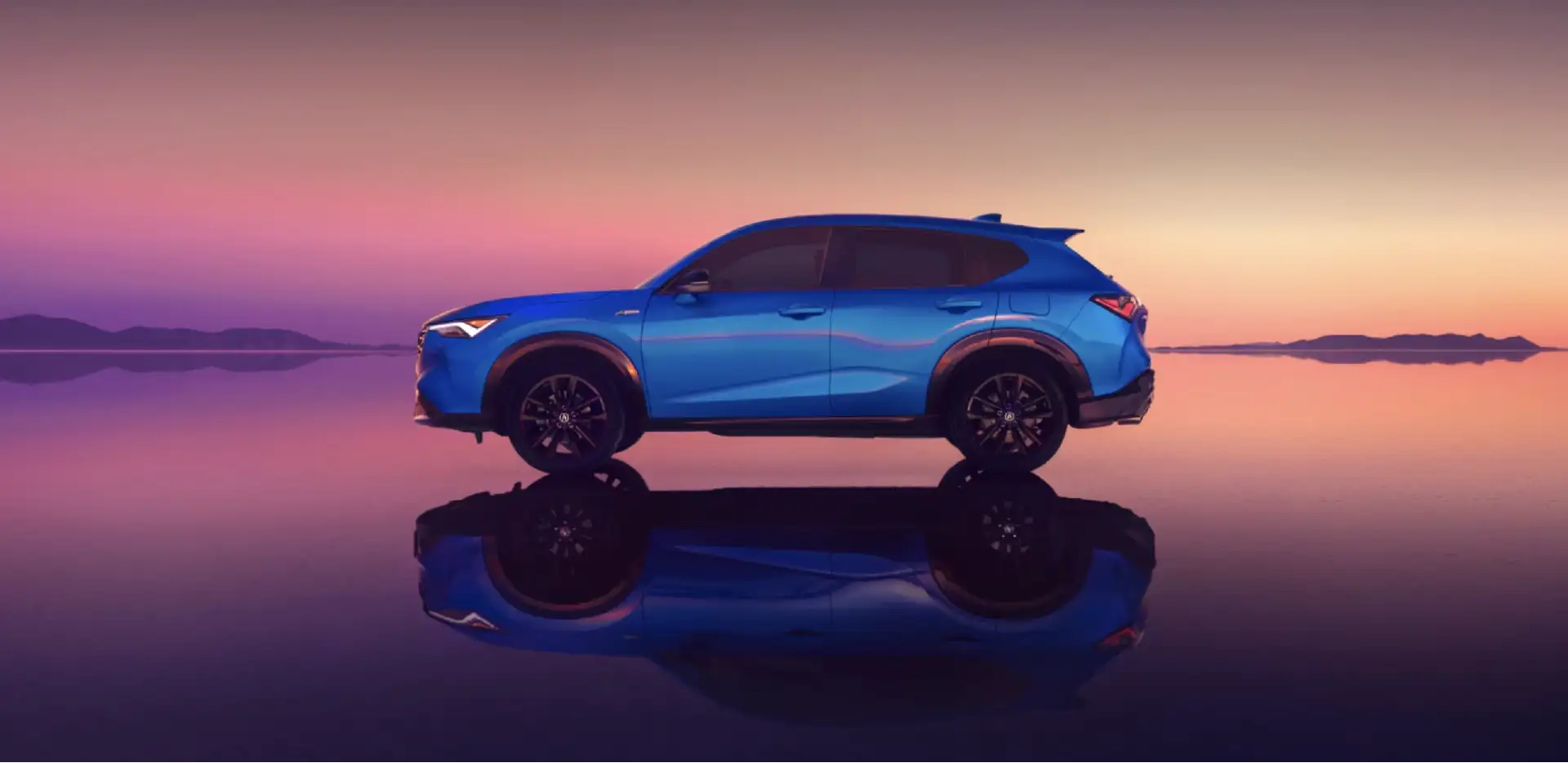 An Acura SUV shown in side profile on a mirror-like flat surface, reflected below against a purple and orange sunset sky