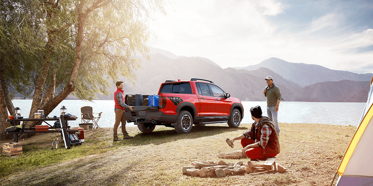 2026 Honda Ridgeline parked along a riverbank with a mountainous backdrop with men getting ready to camp