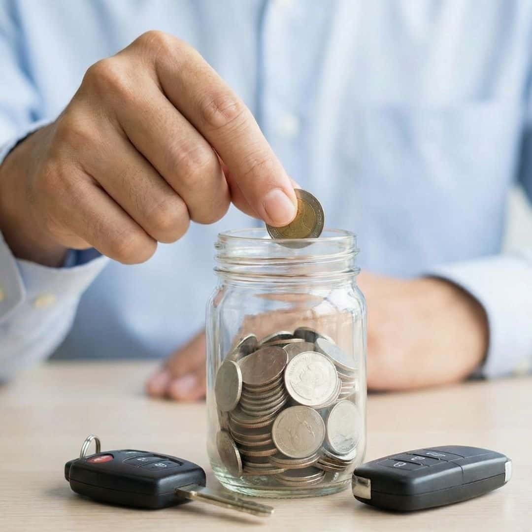 A hand placing coins into a jar next to a car key, representing long-term savings.