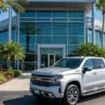 A silver Chevrolet Silverado truck parked in front of a modern dealership building on a sunny day.