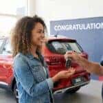 A young woman smiles as she receives keys to a new car at a dealership.