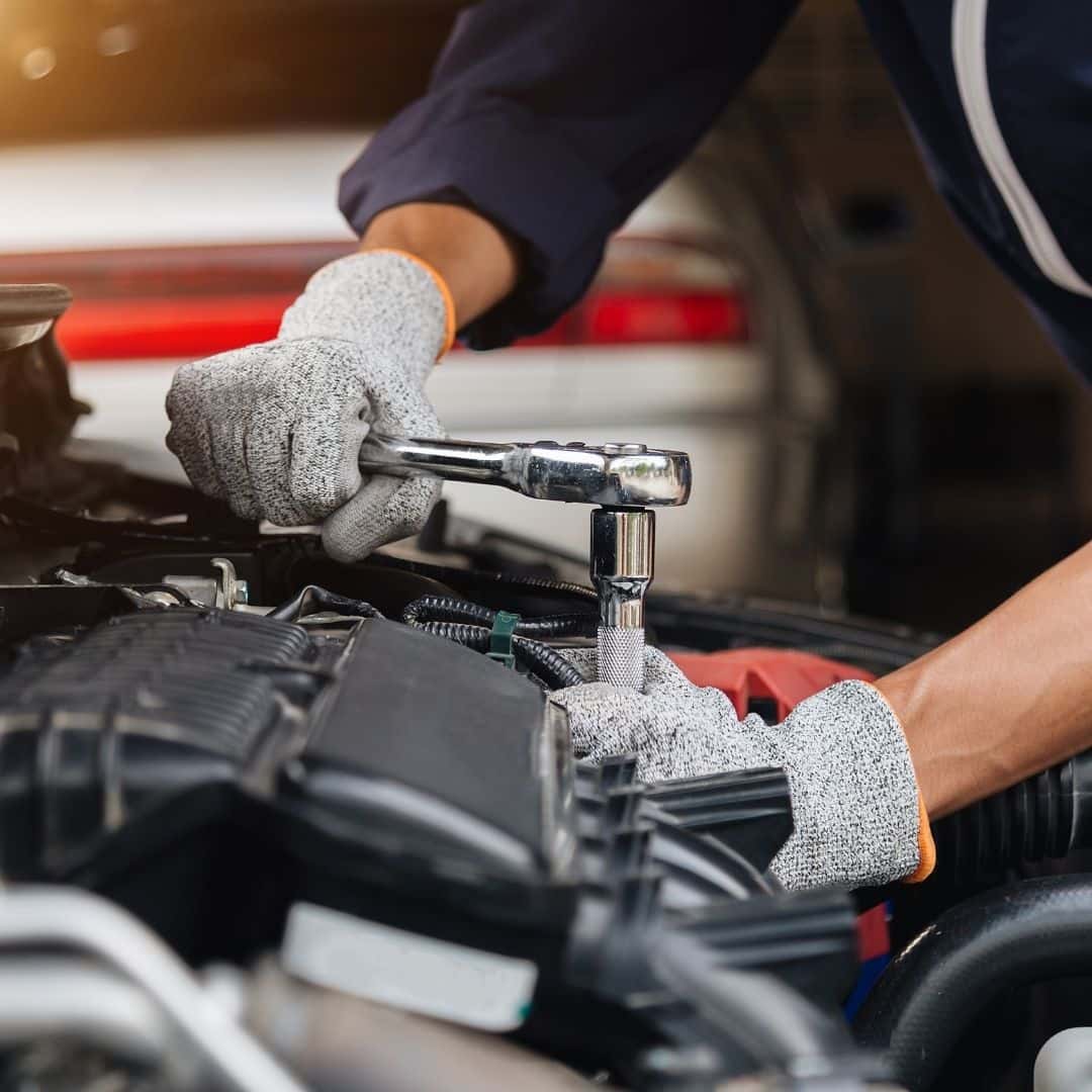 A mechanic in a service bay inspects the engine of a car while a man in business casual attire watches.