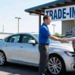 A car salesperson shakes hands with a couple next to a car at a dealership, with a "Trade-In Center" sign visible in the background.