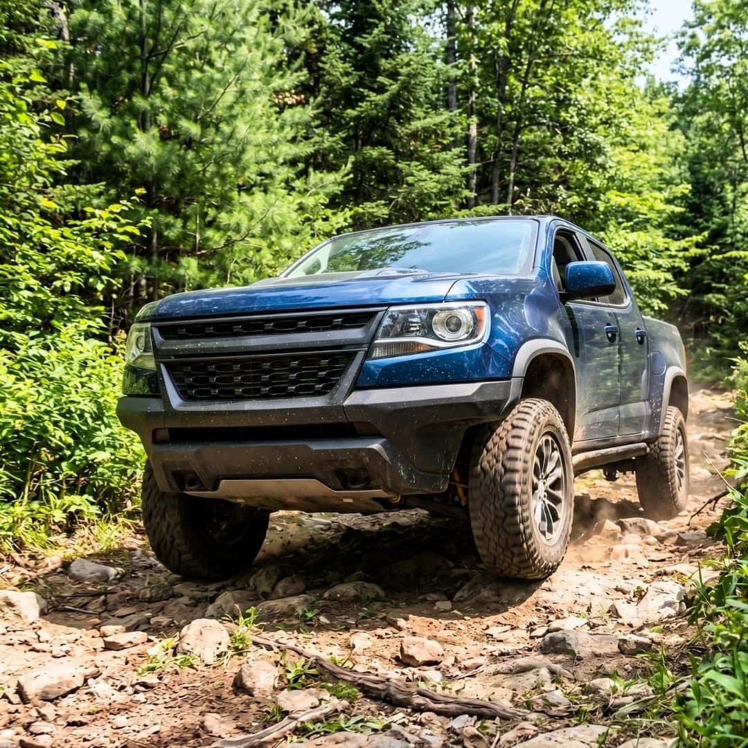 A Chevy Colorado truck driving over a rugged, rocky off-road trail surrounded by trees.