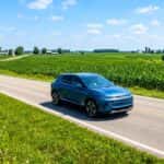 A modern Chevrolet Equinox EV driving down a scenic rural Indiana highway surrounded by green fields under a bright summer sky.
