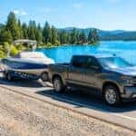 A modern Chevy Silverado 1500 towing a premium boat on a scenic lakeside road during summer.