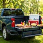 A modern Chevy Silverado truck with its Multi-Flex Tailgate open, serving as a food buffet station during a sunny summer outdoor barbecue party.