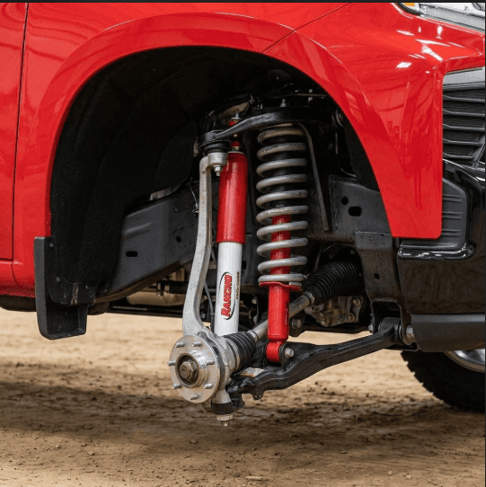 A detailed photograph focuses on the front suspension of a red Z71 truck, highlighting a Rancho shock absorber and coil spring against a clean dirt workshop floor.