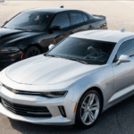 A silver Chevrolet Camaro and a black Dodge Charger are parked side-by-side on asphalt under dynamic daylight.