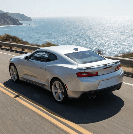 A sleek silver Chevrolet Camaro drives along a sunlit coastal highway, displaying its athletic coupe profile against the ocean background.