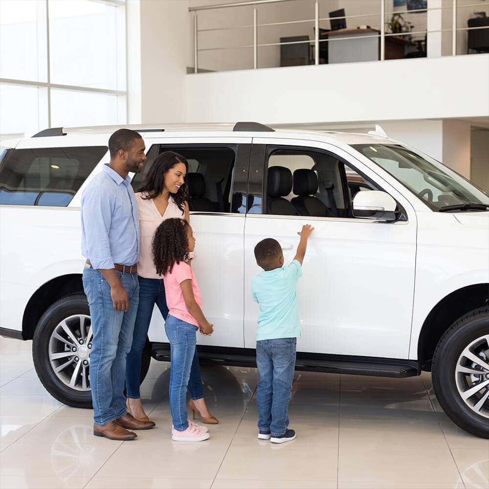 A diverse family looking at the interior of a large new white SUV in a brightly lit showroom.