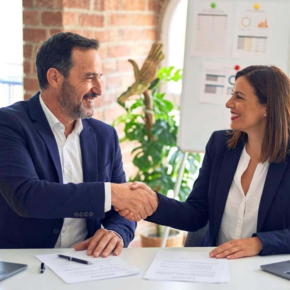 Two people smiling and shaking hands over papers on a desk.