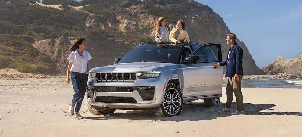 A family standing in and beside a 2026 Jeep Grand Cherokee SUV parked on a beach