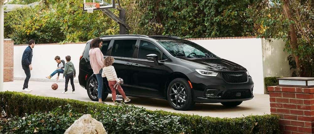 A family playing basketball beside a black 2026 Chrysler Pacifica van parked in their driveway
