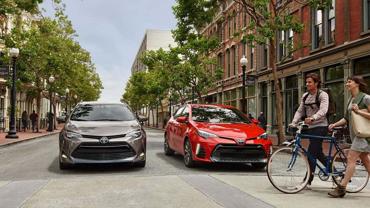 Couple walks in front of red and brown Toyota Corollas at crosswalk