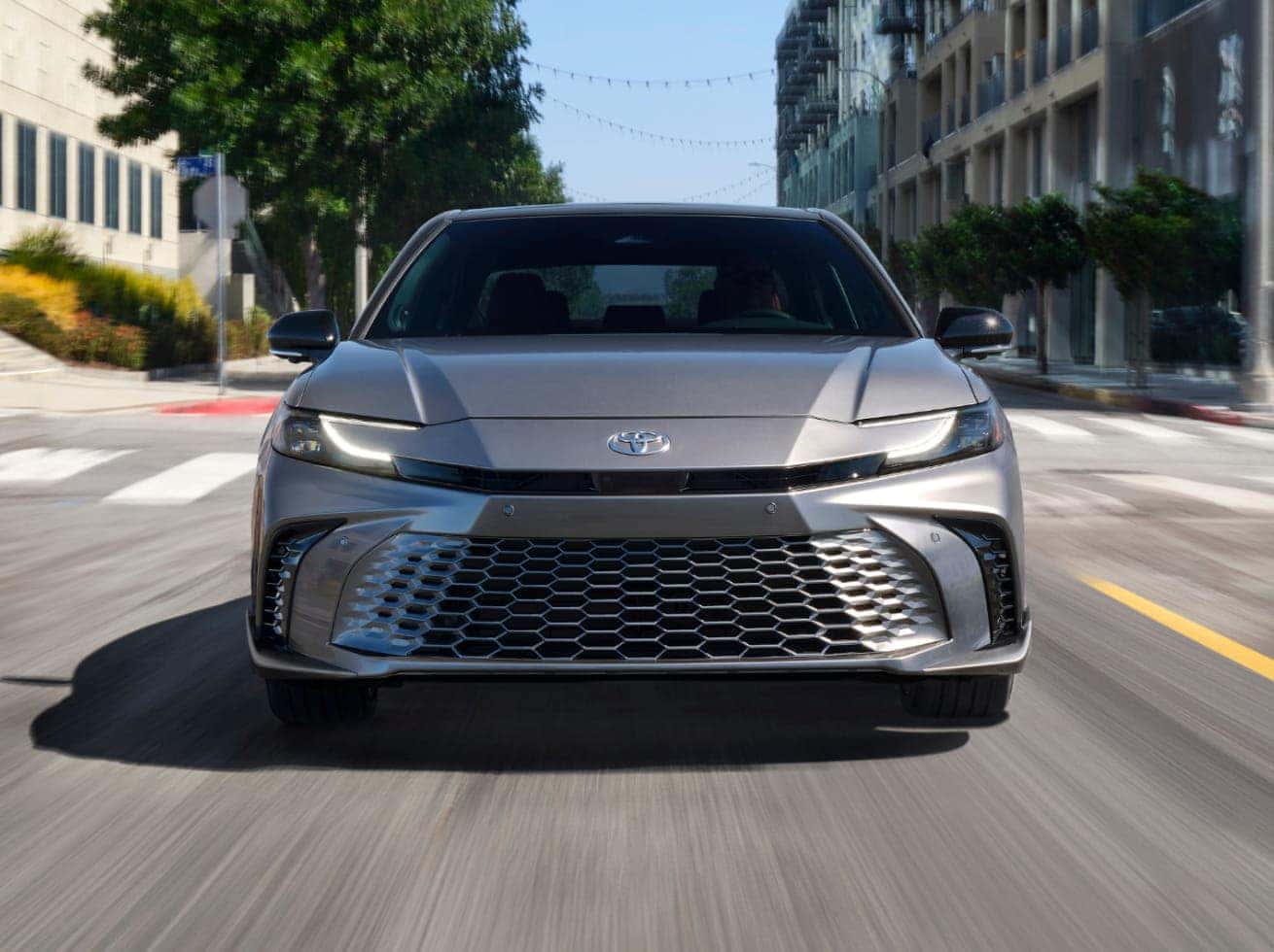 Front view of a 2026 Toyota Camry sedan driving on a city street. Urban background.