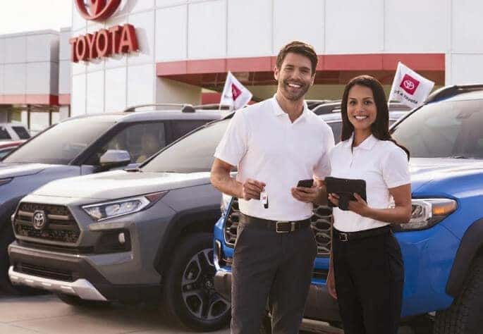 Two Toyota sales representatives smile outside a Toyota dealership on a sunny day.