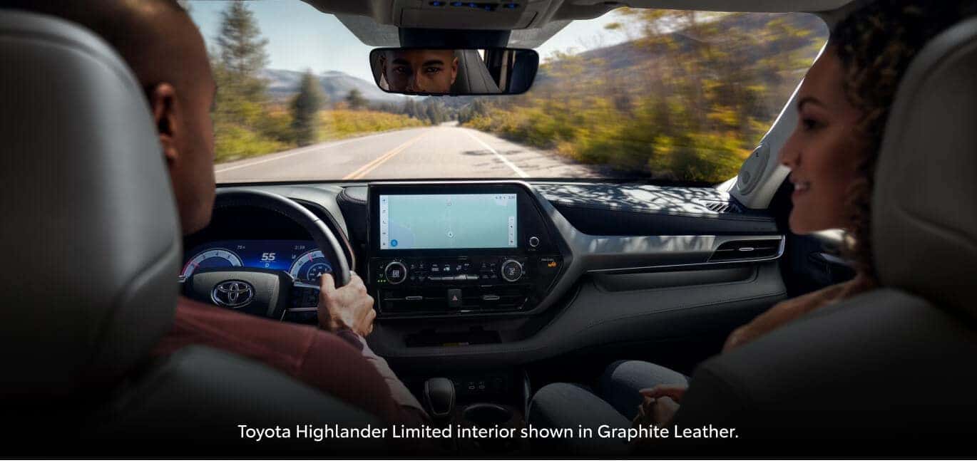 Interior view of a couple driving a gray Toyota Highlander Limited down an open road lined with trees.