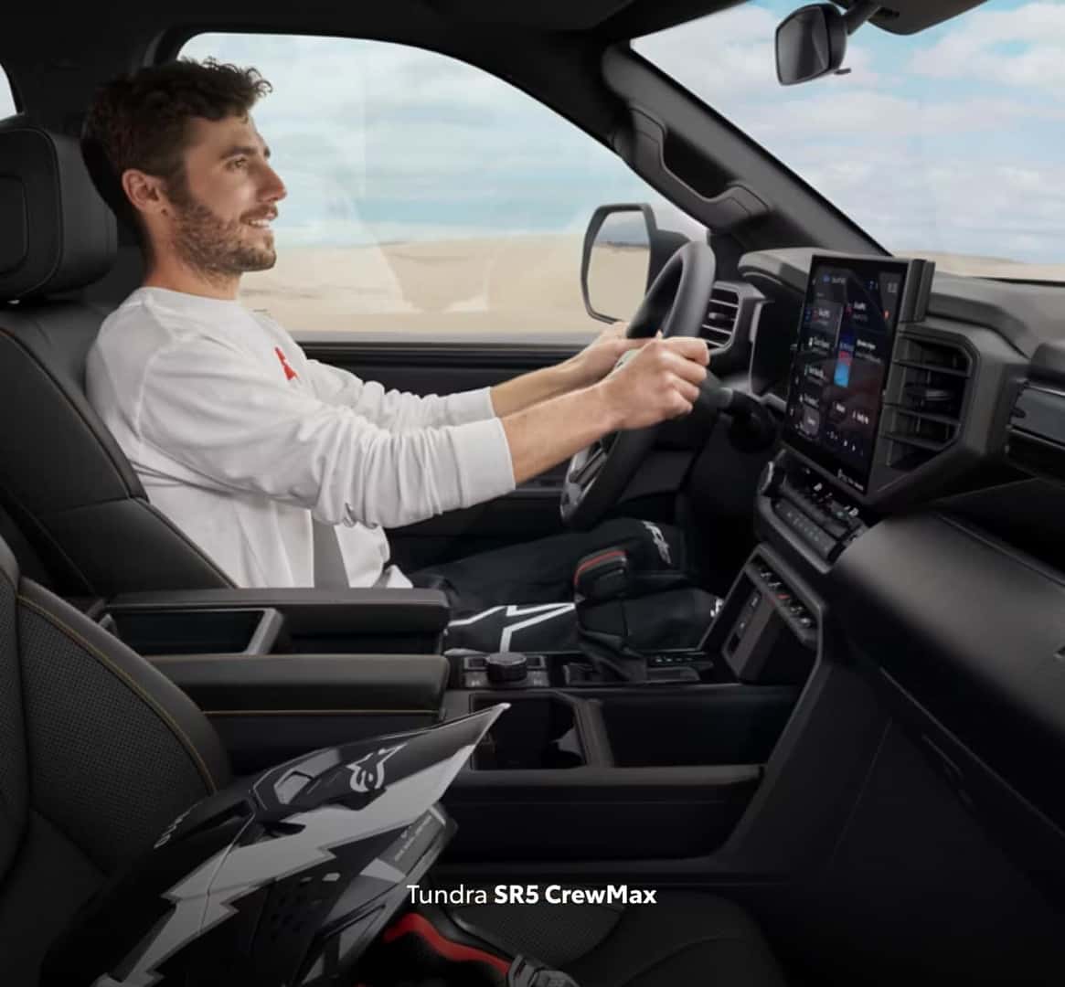 Driver behind the wheel of a Toyota Tundra SR5 CrewMax, with the touchscreen display and spacious cabin visible.