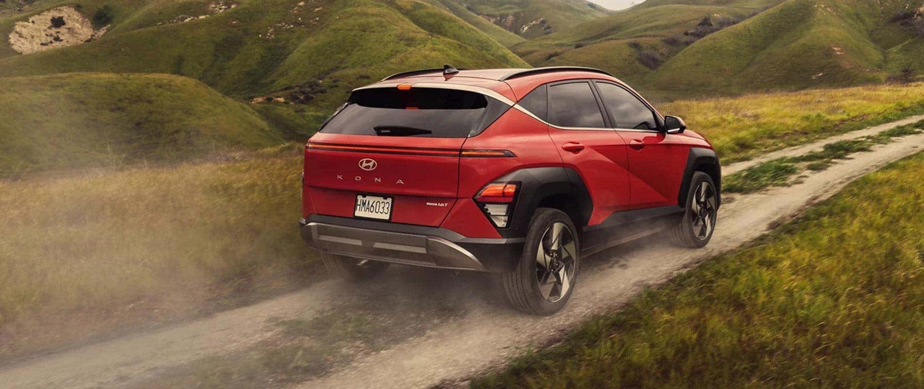 A red 2025 Hyundai Kona driving over a dirt road through a hilly field.