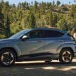 A light blue Hyundai KONA Electric by a lake in a forest, with a woman leaning against its trunk.