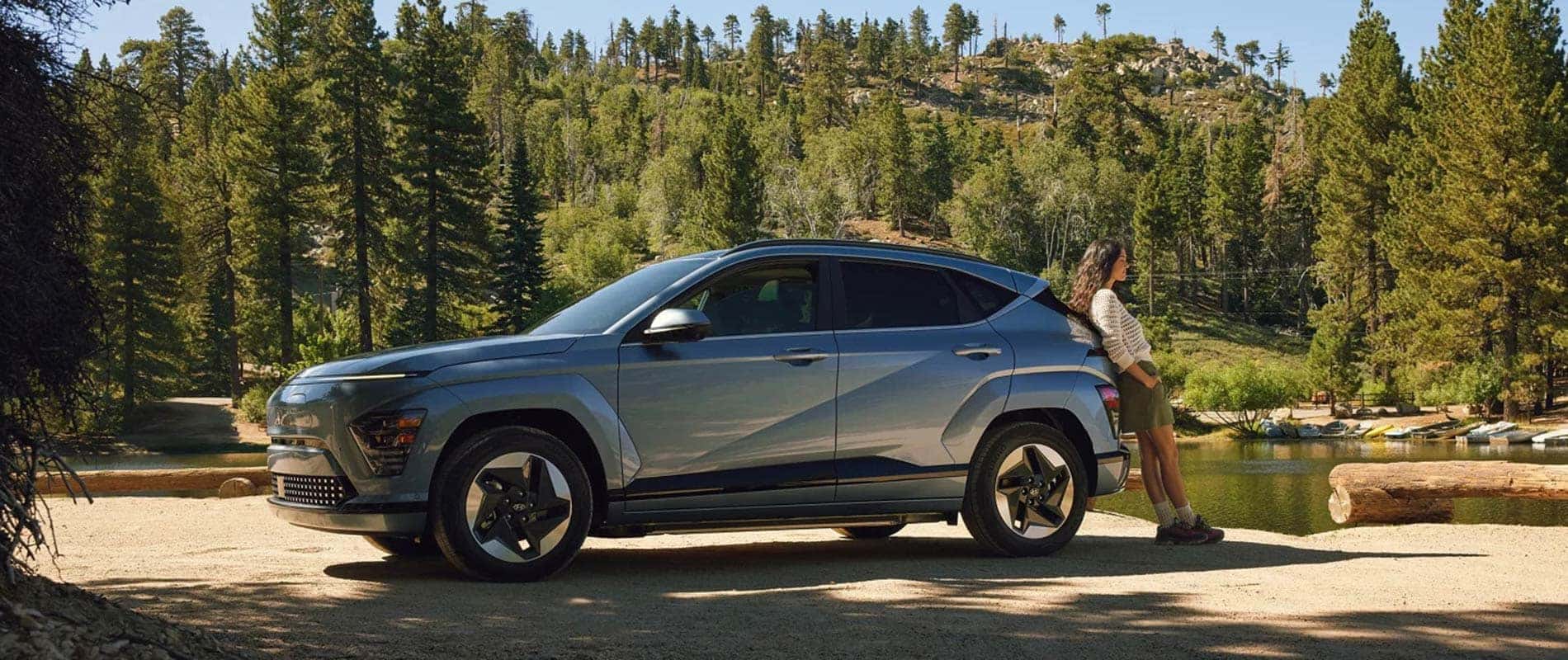 A light blue Hyundai KONA Electric by a lake in a forest, with a woman leaning against its trunk.