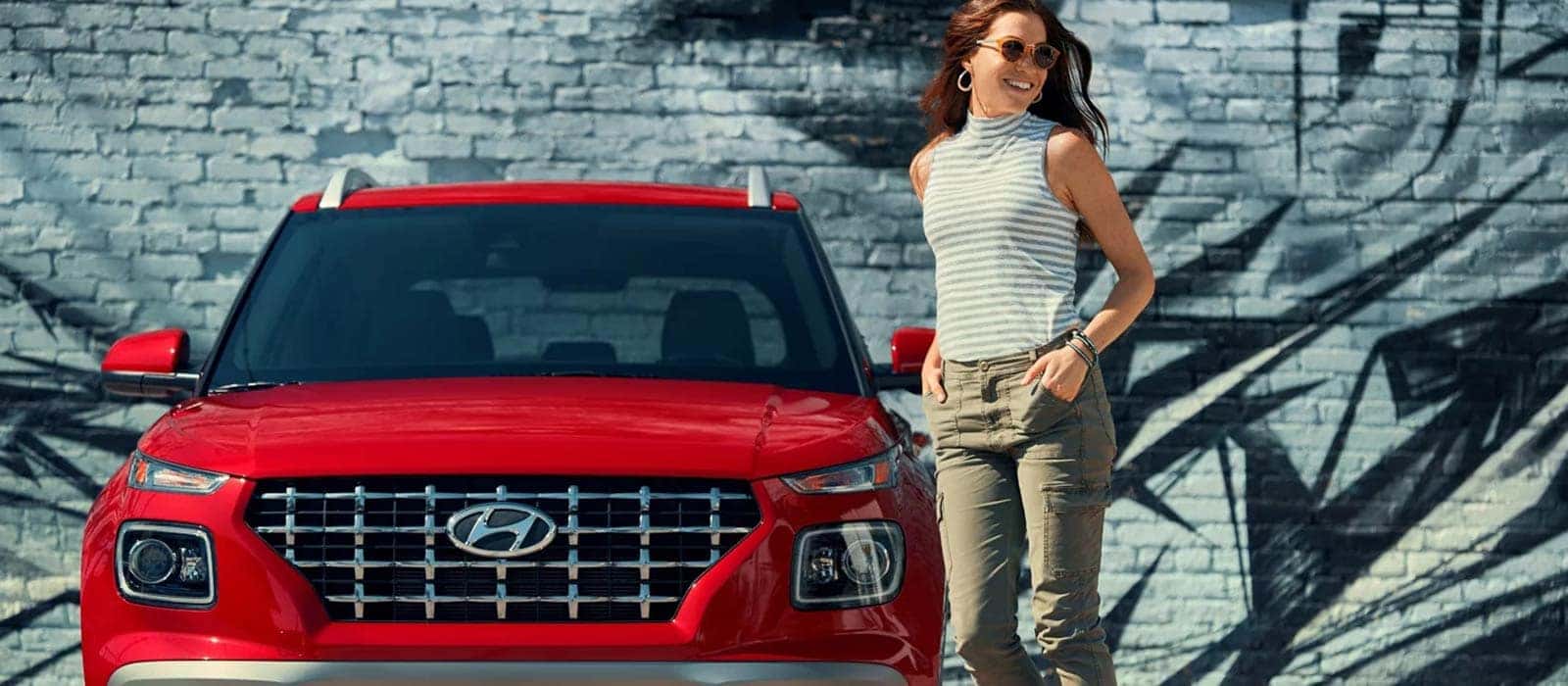A front view of a red Hyundai Venue with a woman standing next to it in front of a graffitied brick wall.