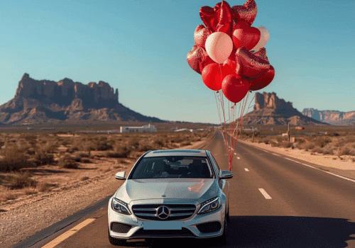 Silver Mercedes-Benz sedan driving on desert highway with red heart balloons tied to roof.