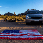 SUV parked in the desert beside an American flag laid out on the ground with mountains in the background.