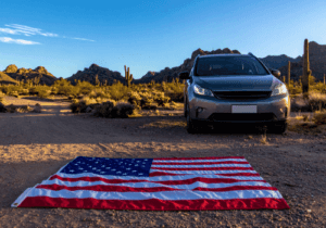SUV parked in the desert beside an American flag laid out on the ground with mountains in the background.