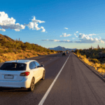 White station wagon driving along a desert highway surrounded by cacti, mountains, and clear summer skies.