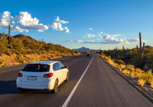 White station wagon driving along a desert highway surrounded by cacti, mountains, and clear summer skies.