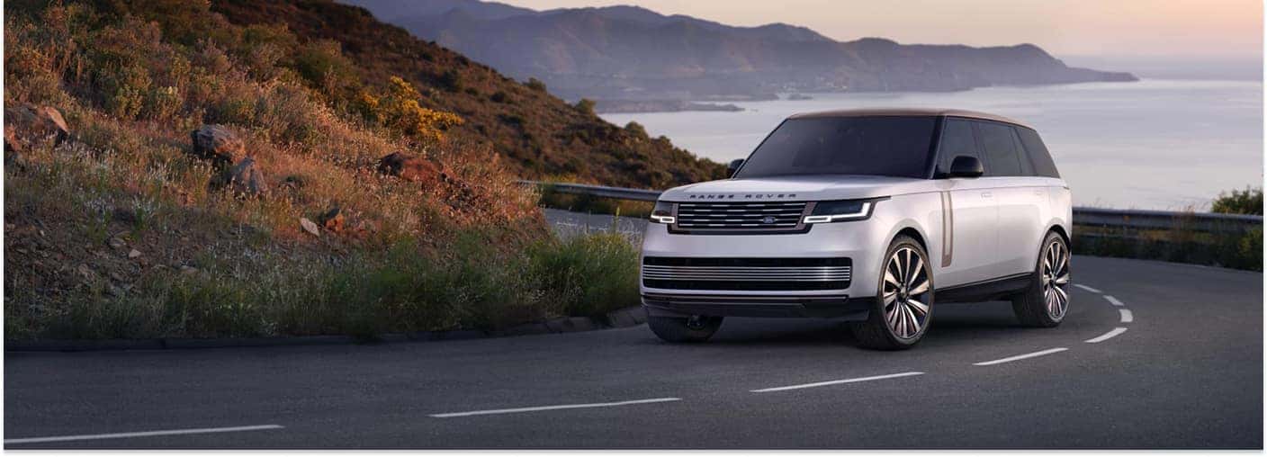 Front three-quarter view of a white 2025 Range Rover SUV driving on a coastal road at sunset.