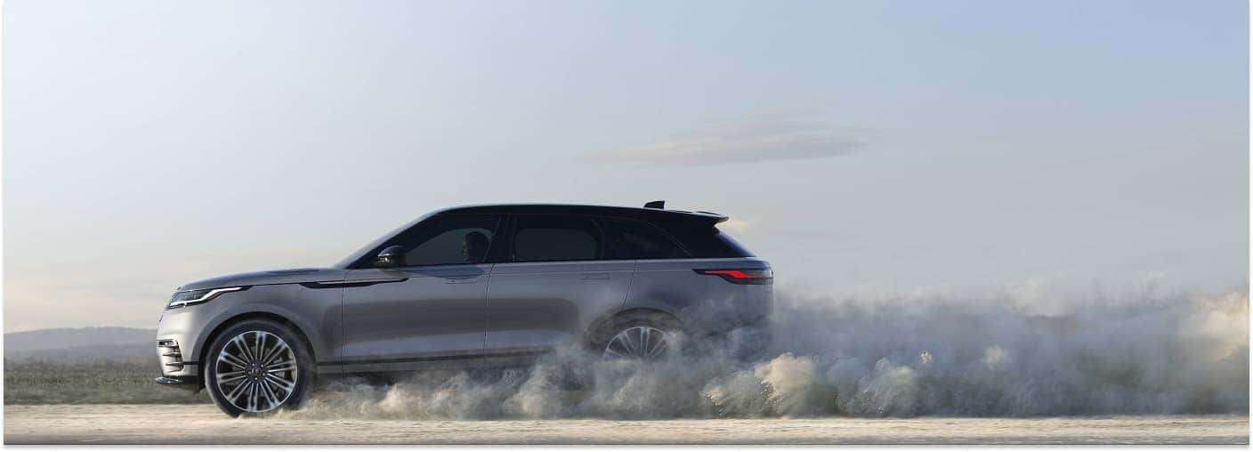 Side profile of a silver 2025 Range Rover Velar driving on a dirt road and kicking up dust.
