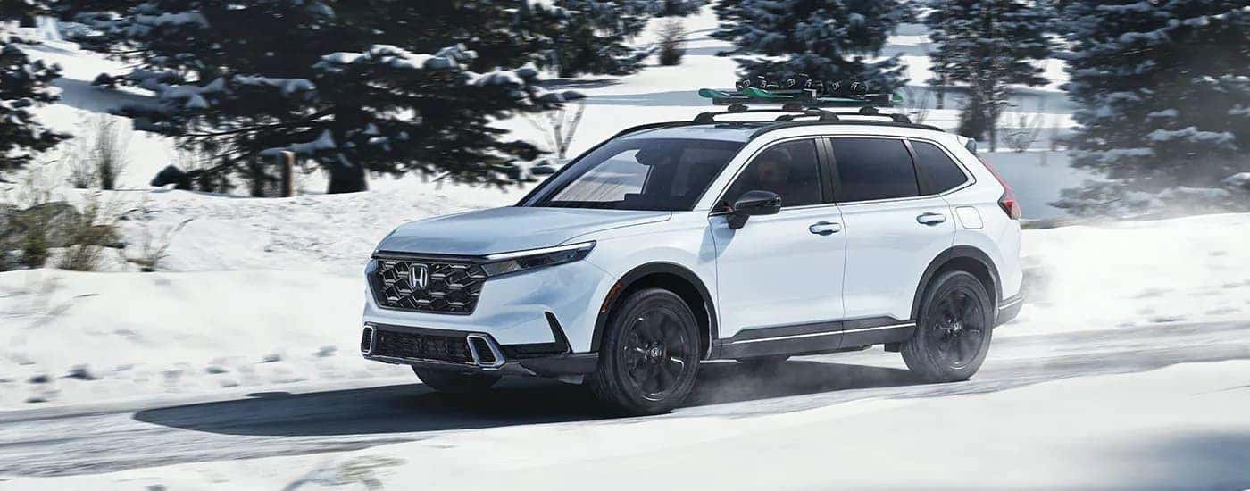 A white 2025 Honda CR-V Sport Touring Hybrid driving down a snowy road.