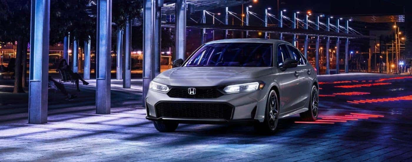 A silver 2025 Honda Civic Hybrid Sport driving down a city street at night