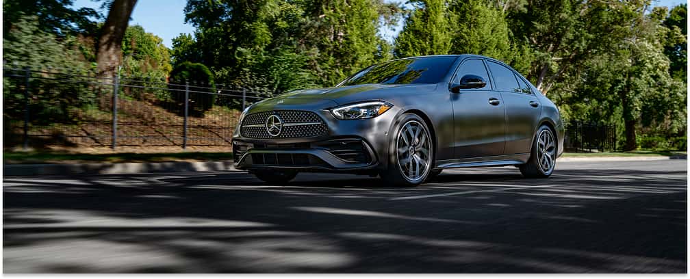 Matte gray 2026 Mercedes-Benz C-Class sedan driving on a tree-lined street.