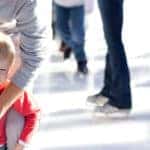 Child ice skating with guardian at a crowded rink on a sunny day
