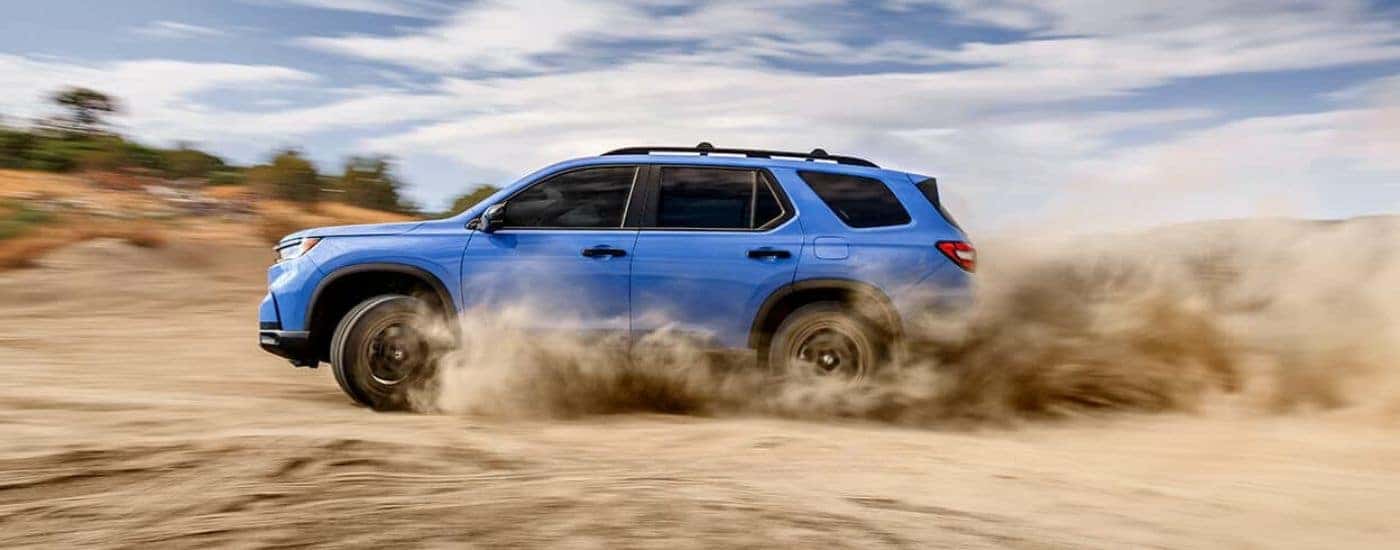 A popular Honda Pilot for sale near Fort Worth, a blue 2024 Honda Pilot TrailSport, is shown driving off-road.