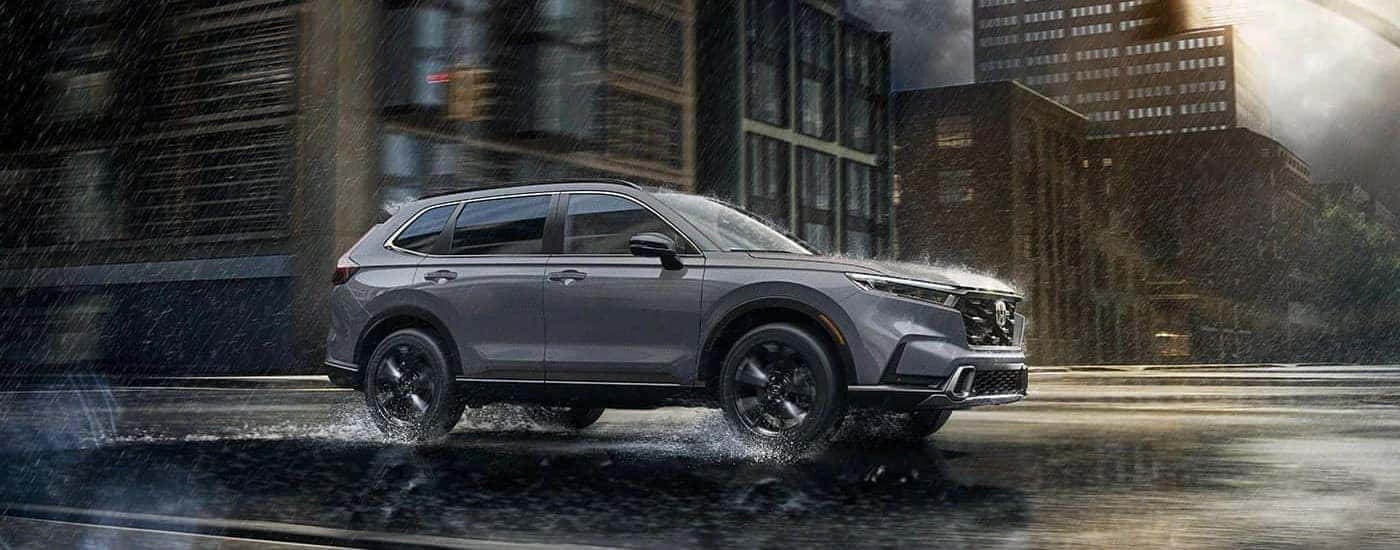 Side angle view of a grey 2023 Honda CR-V Sport Touring Hybrid driving through a city intersection during the rain.