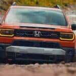 Front grille view of an orange 2026 Honda Passport TrailSport driving in mud.