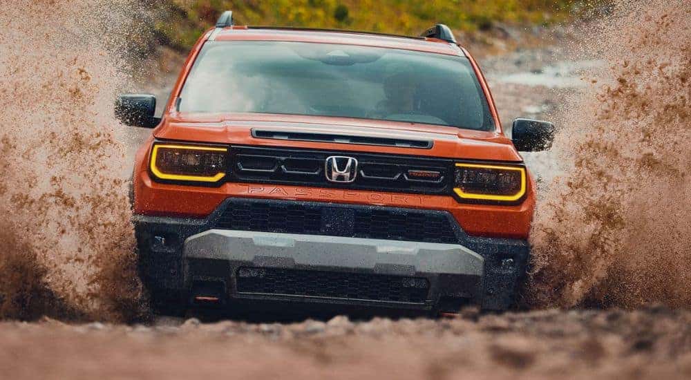 Front grille view of an orange 2026 Honda Passport TrailSport driving in mud.