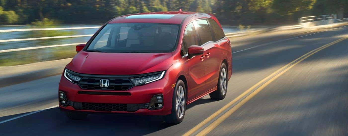 Red 2026 Honda Odyssey driving on a paved road along a lake.