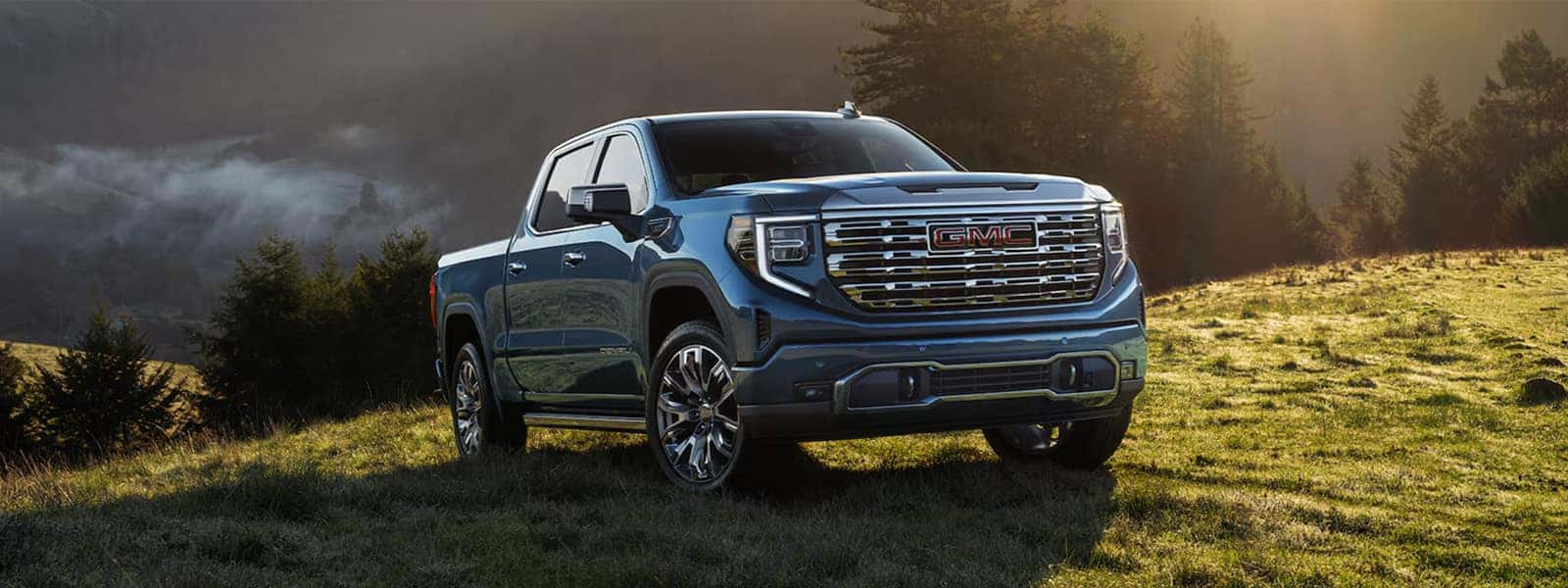 2026 GMC Sierra 1500 pickup truck parked on a grassy hillside at sunrise, showcasing bold grille design and premium exterior styling.