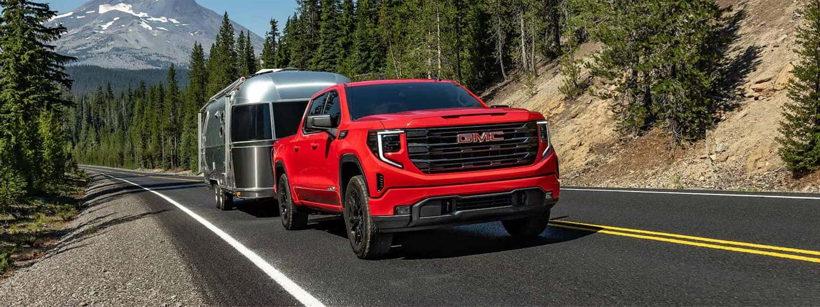 Red 2026 GMC Sierra 1500 towing a silver travel trailer on a mountain highway with pine trees in the background.