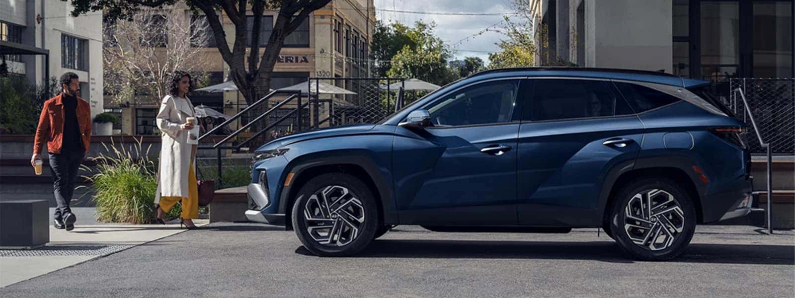 Side profile of a blue Hyundai Tucson Hybrid parked in an urban setting, highlighting sleek exterior design and modern SUV styling.