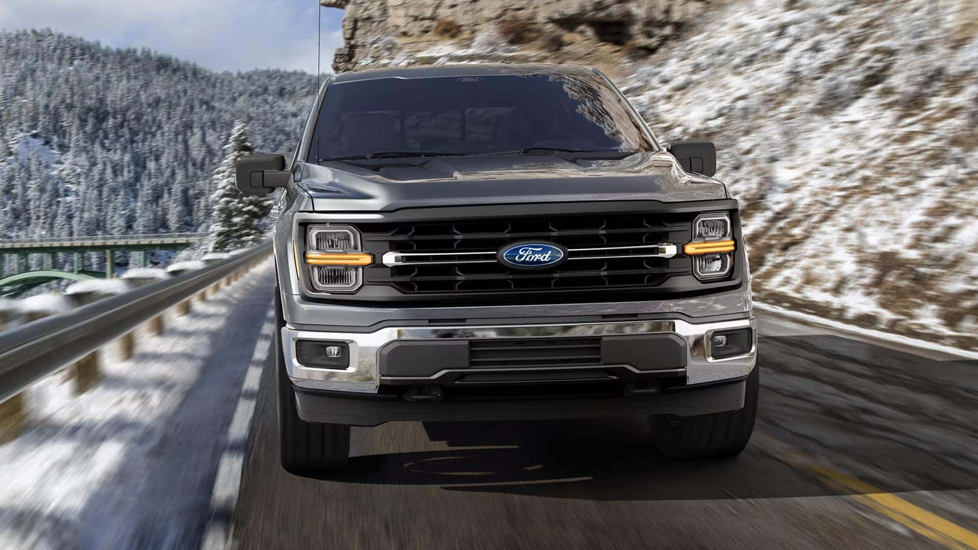 Front view of a 2026 Ford F-150 driving on a mountain road, showcasing bold grille design, LED headlamps, and strong exterior styling.