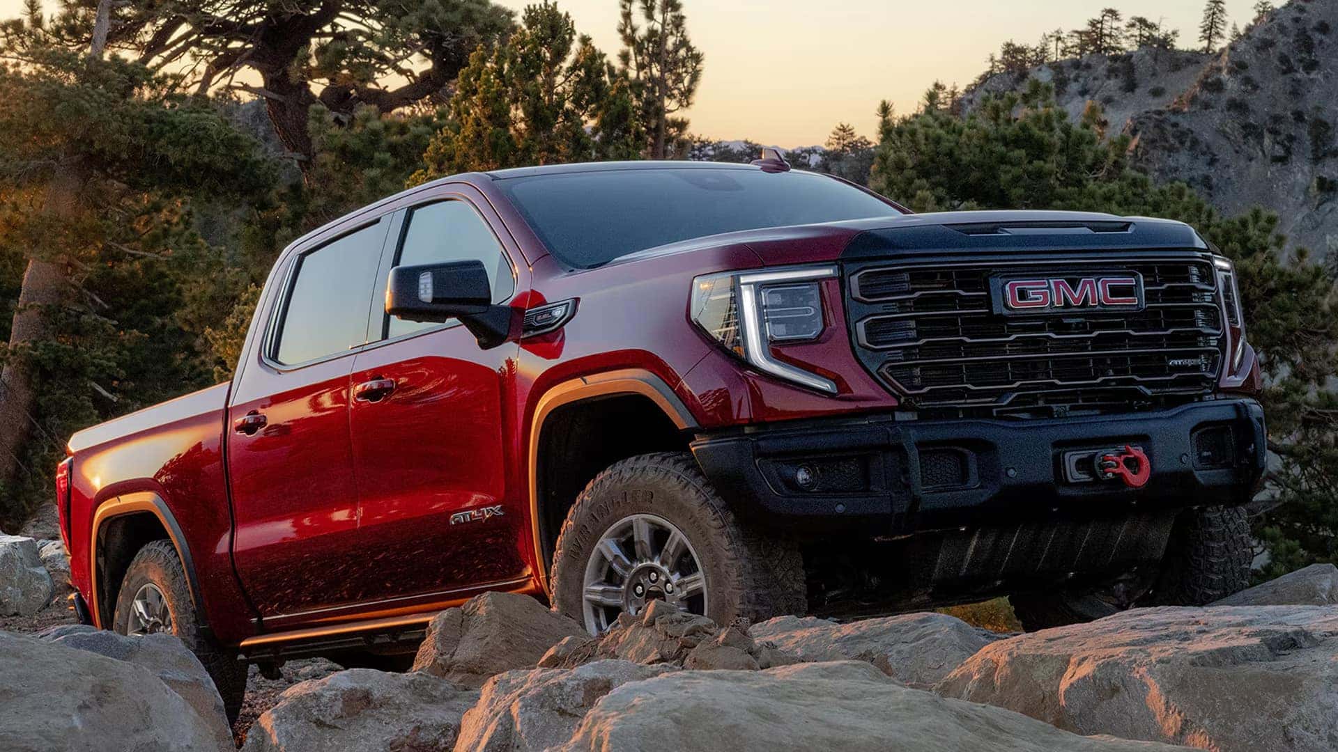 Red 2026 GMC Sierra 1500 AT4X driving over rocky terrain, highlighting off-road capability, lifted stance, and rugged front bumper.
