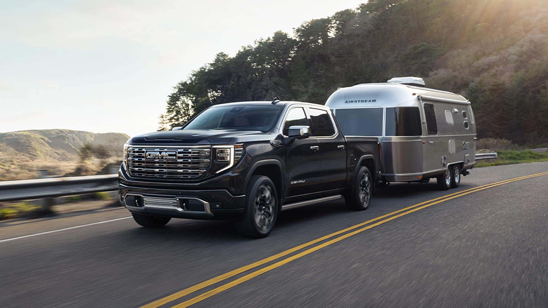 Black 2026 GMC Sierra 1500 towing an Airstream travel trailer on a coastal road in late afternoon light.
