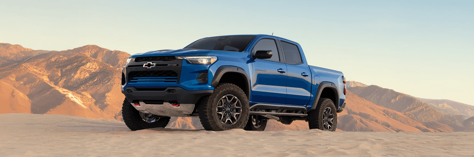 Blue Chevy Colorado parked on sand dunes with mountain backdrop